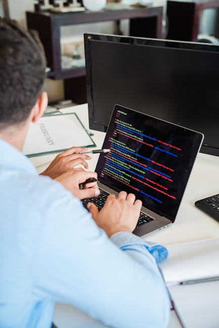 A person working on a laptop that displays a screen of computer code with colorful syntax highlighting, seated at a desk in an indoor setting. The desk holds a closed notebook labeled 'REFERENCES', a 